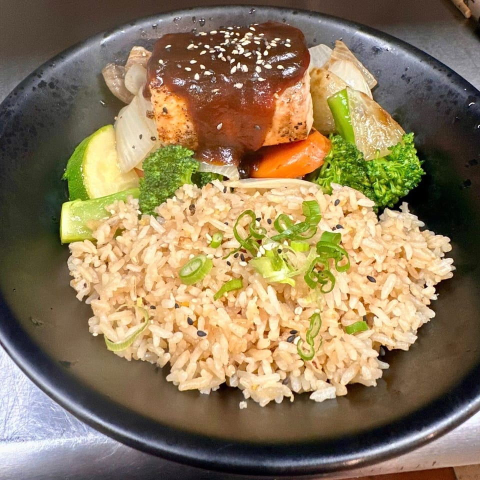 Asian-fusion salmon with dark sauce, sesame seeds, mixed vegetables, and fried rice in a black bowl.