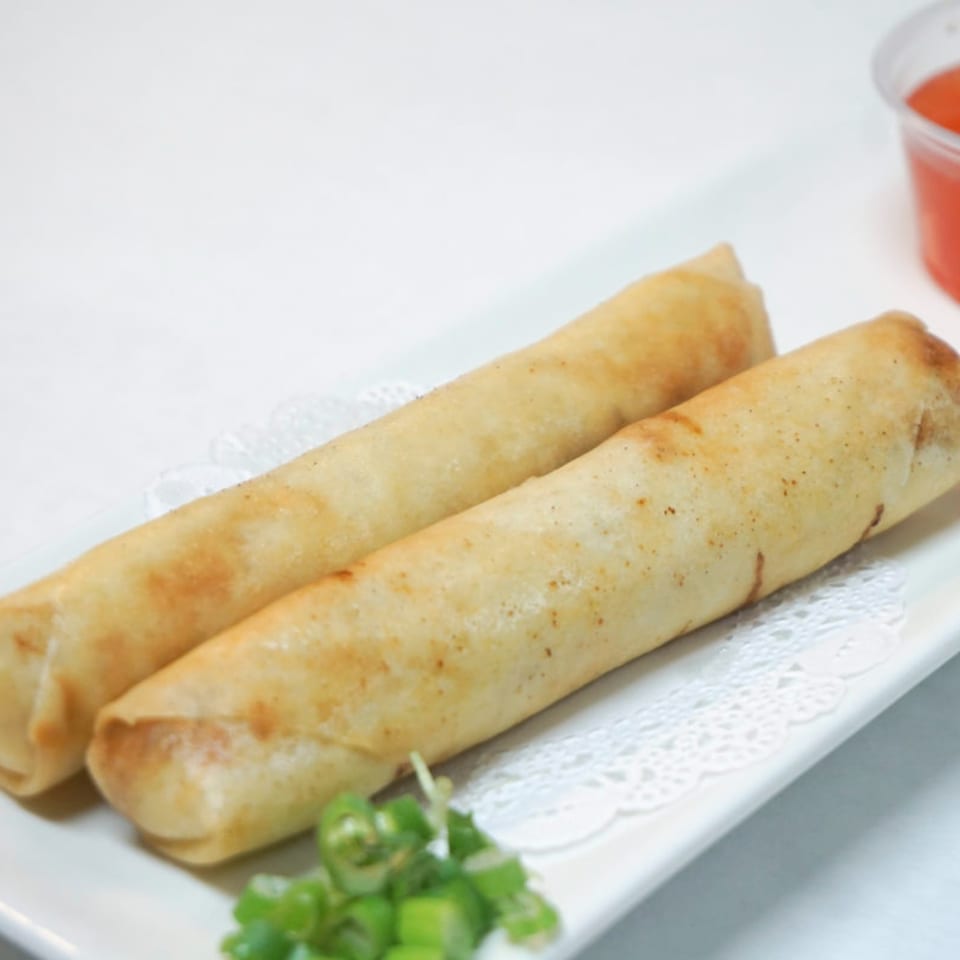 Two golden-brown spring rolls on a white plate with a side of sweet chili sauce and green onions.