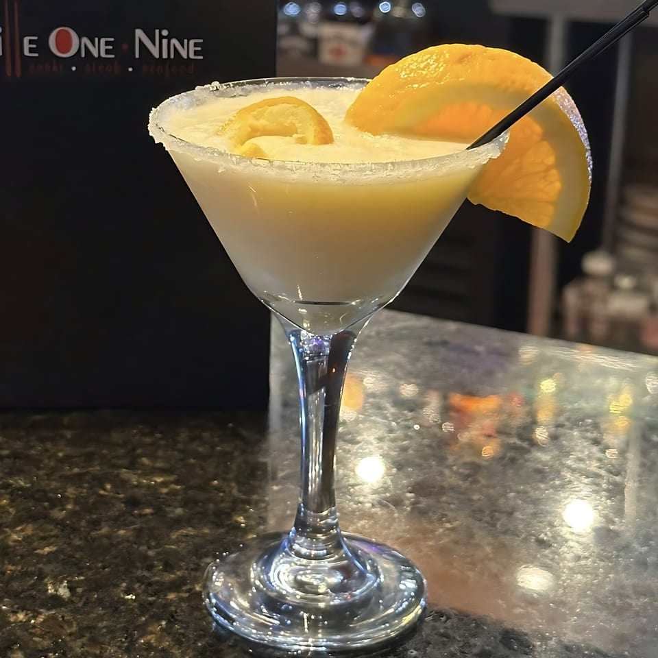 A Grille One Nine cocktail, a martini glass with a sugared rim, orange slices, and a black straw.