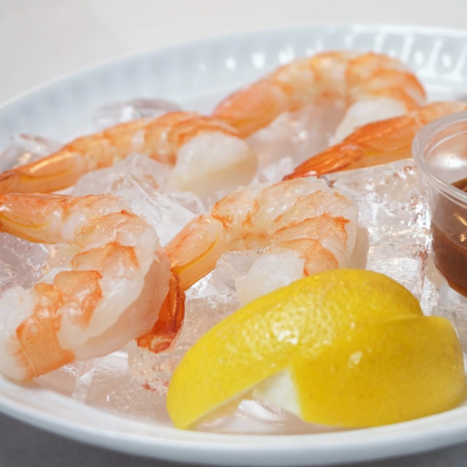 Shrimp cocktail on ice with a lemon wedge and dipping sauce on a white plate.
